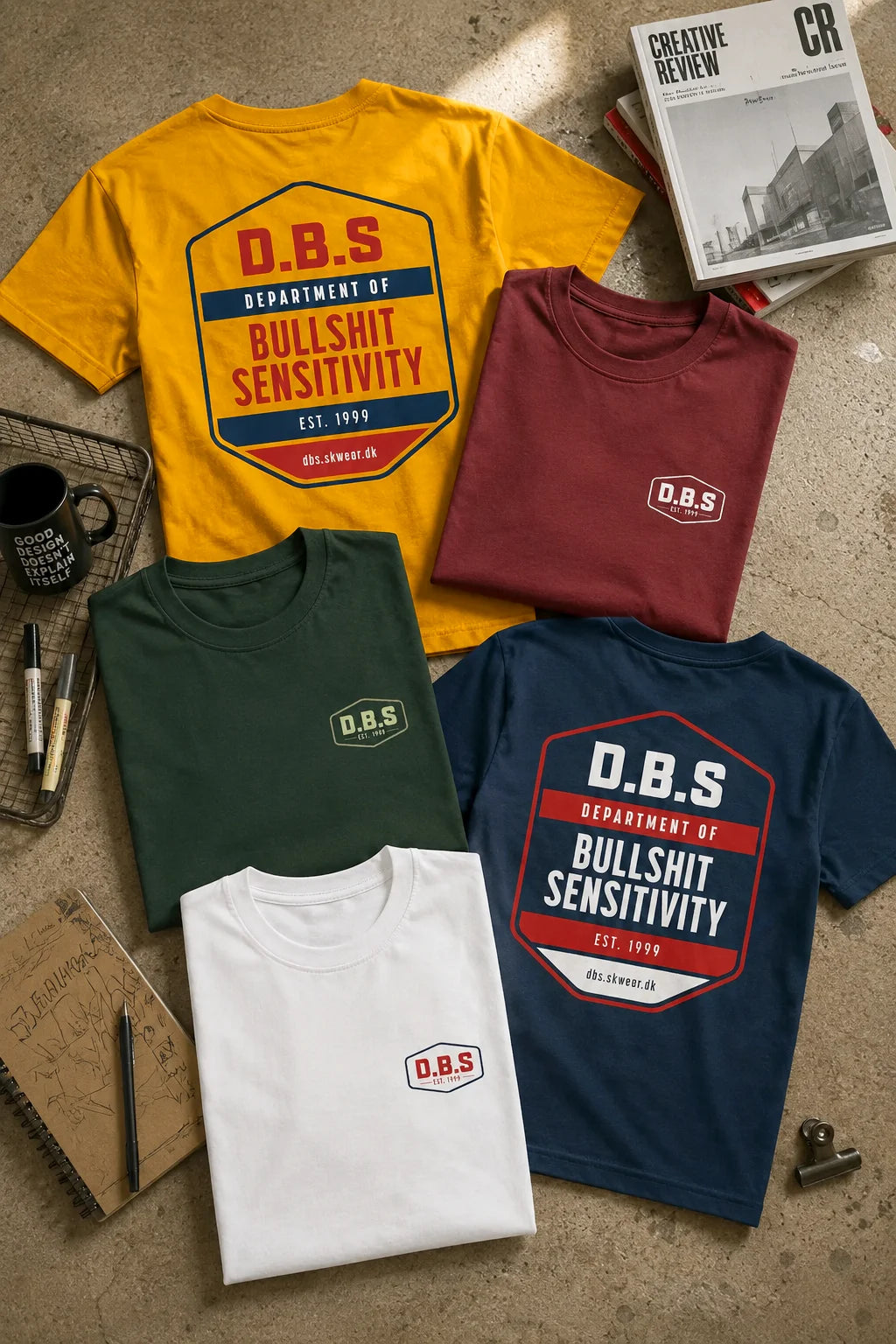 Five t-shirts with D.B.S. logo on a textured surface with books and a mug.